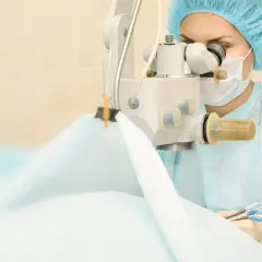 Chirurg w masce i czepku wykonuje operację zaćmy, używając mikroskopu i precyzyjnych narzędzi.