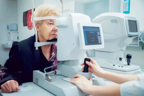 Starsza pani przechodzi badanie pola widzenia u okulisty. Aparatura diagnostyczna jest nowoczesna.