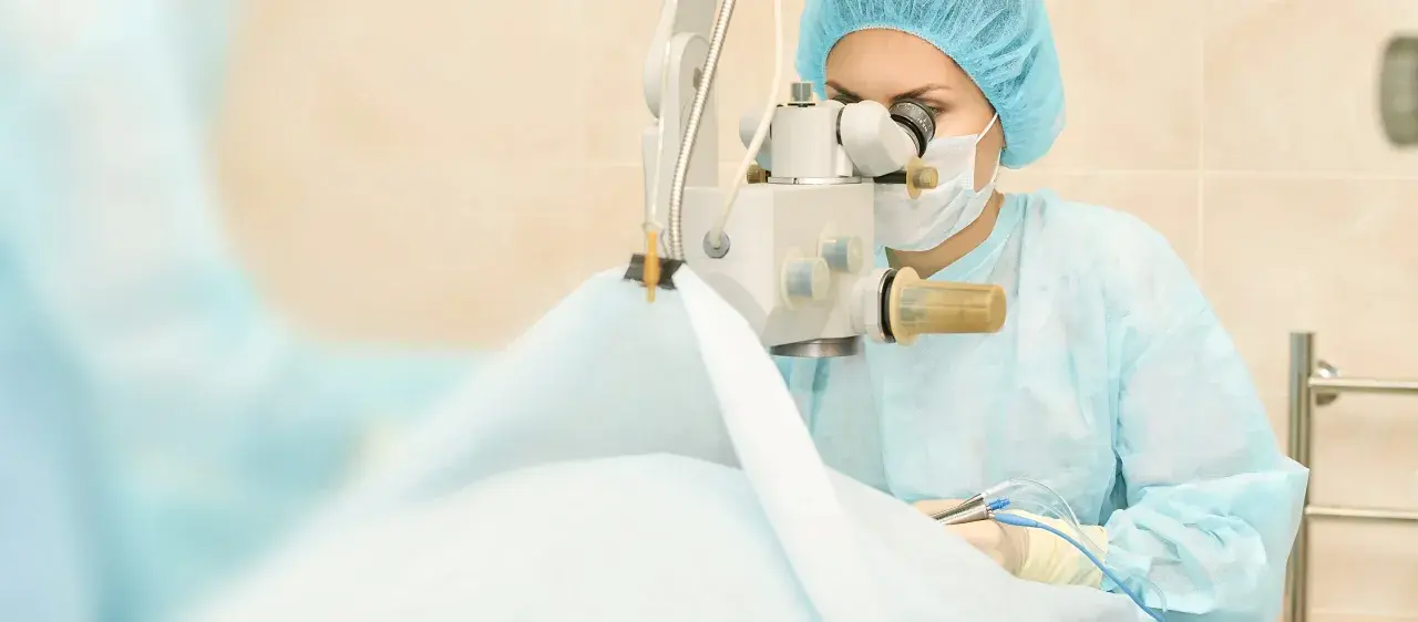 Chirurg w masce i czepku wykonuje operację zaćmy, używając mikroskopu i precyzyjnych narzędzi.
